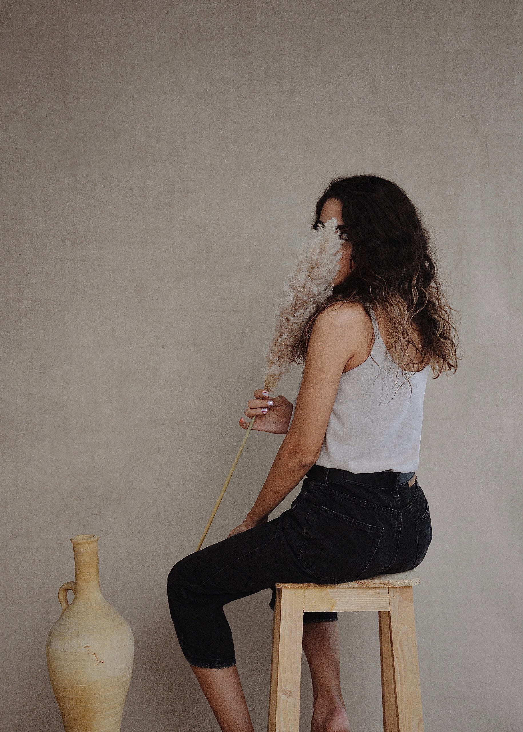 Mujer joven sentada en un banco tapando su rostro con una hoja de Césped de caña artificial y a lado un jarrón de arcilla que se encuentra en el piso